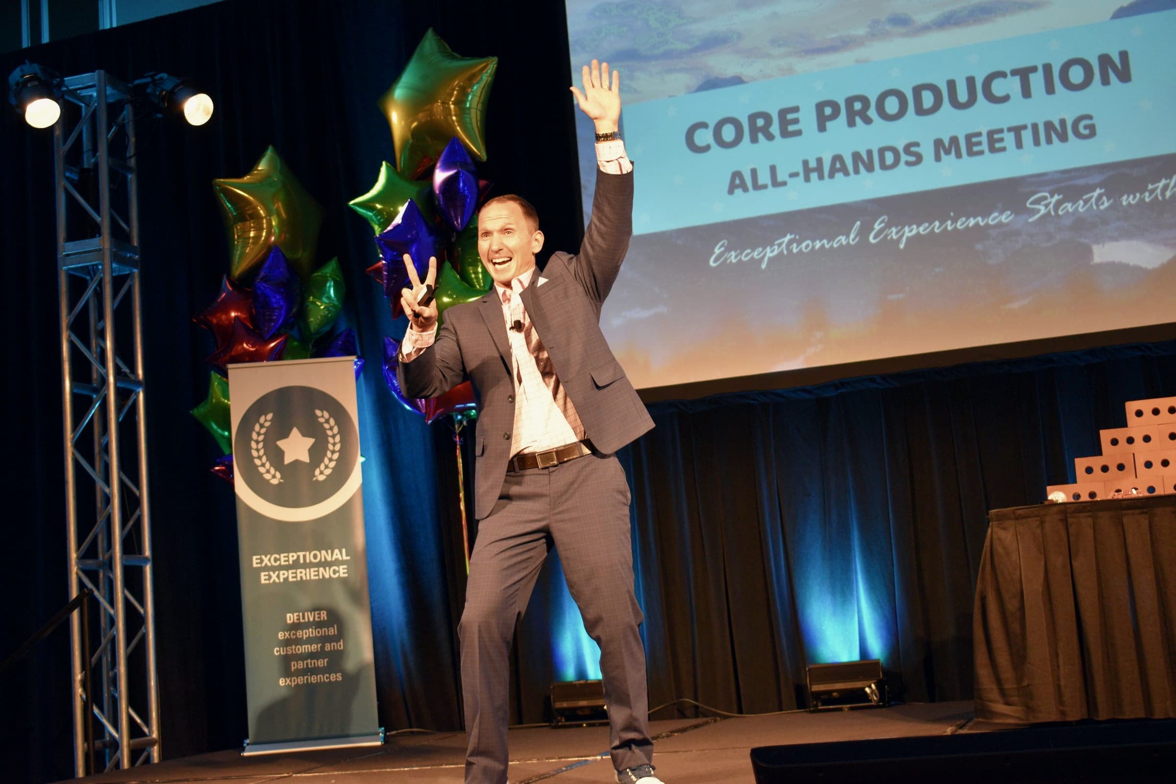Kevin on stage at a company all-hands meeting, arms raised, with balloons and a large screen
