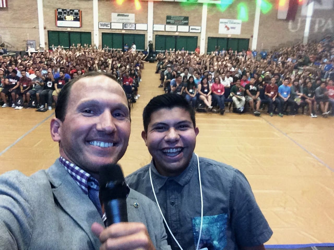 Kevin taking a selfie with a student in front of a packed gymnasium audience