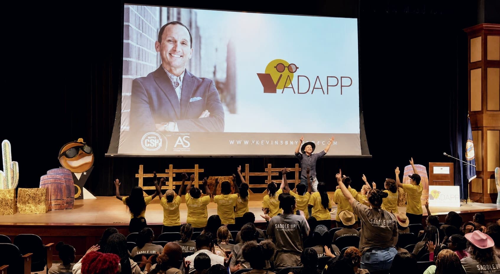 Kevin on stage beside a large screen as a youth audience waves back